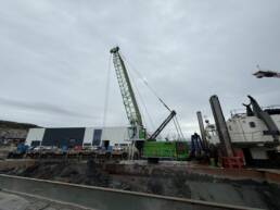 Green crane towering over a muddy harbor construction site with a large building in the background