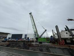 Green crane towering over a muddy harbor construction site with a large building in the background