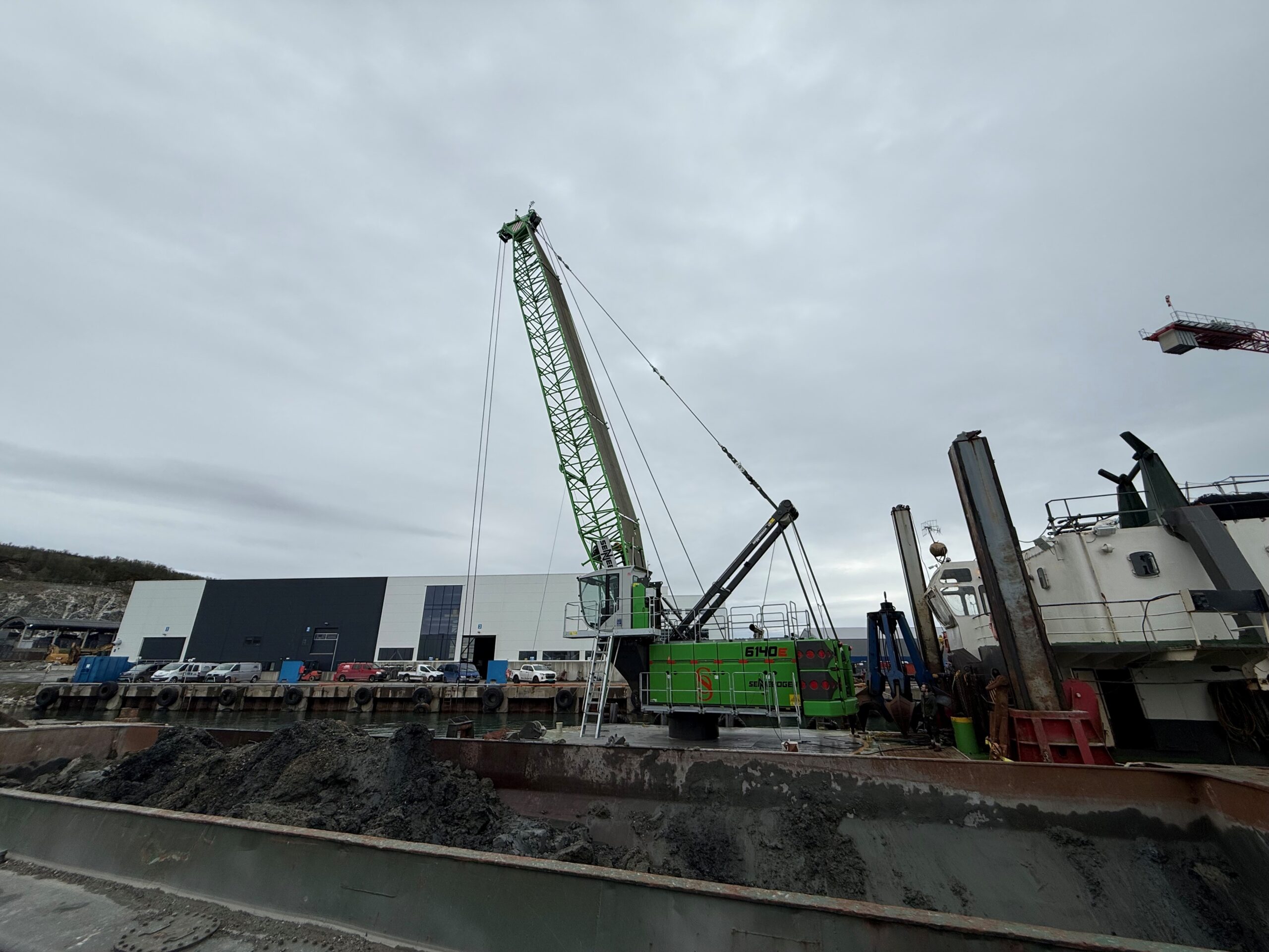 Green crane towering over a muddy harbor construction site with a large building in the background