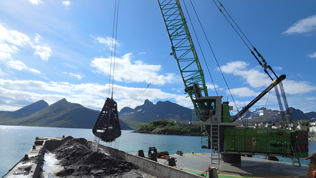 Green heavy-lift crane by the water, with a bucket suspended from cables, mountains and a blue sky in the background.